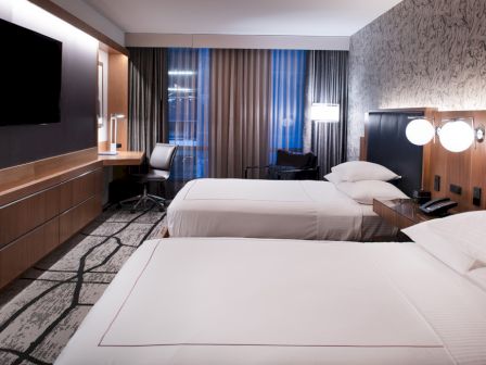 The image shows a modern hotel room with two beds, a wall-mounted TV, a desk, and a window with curtains.