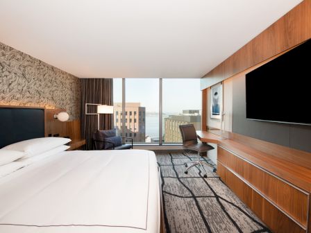 A modern hotel room with a large bed, desk, chair, and television, featuring a window with a cityscape view.