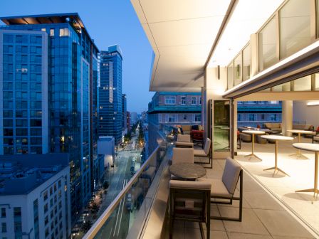 A modern rooftop balcony at dusk, featuring seating with tables, overlooking a cityscape of tall buildings and illuminated streets.