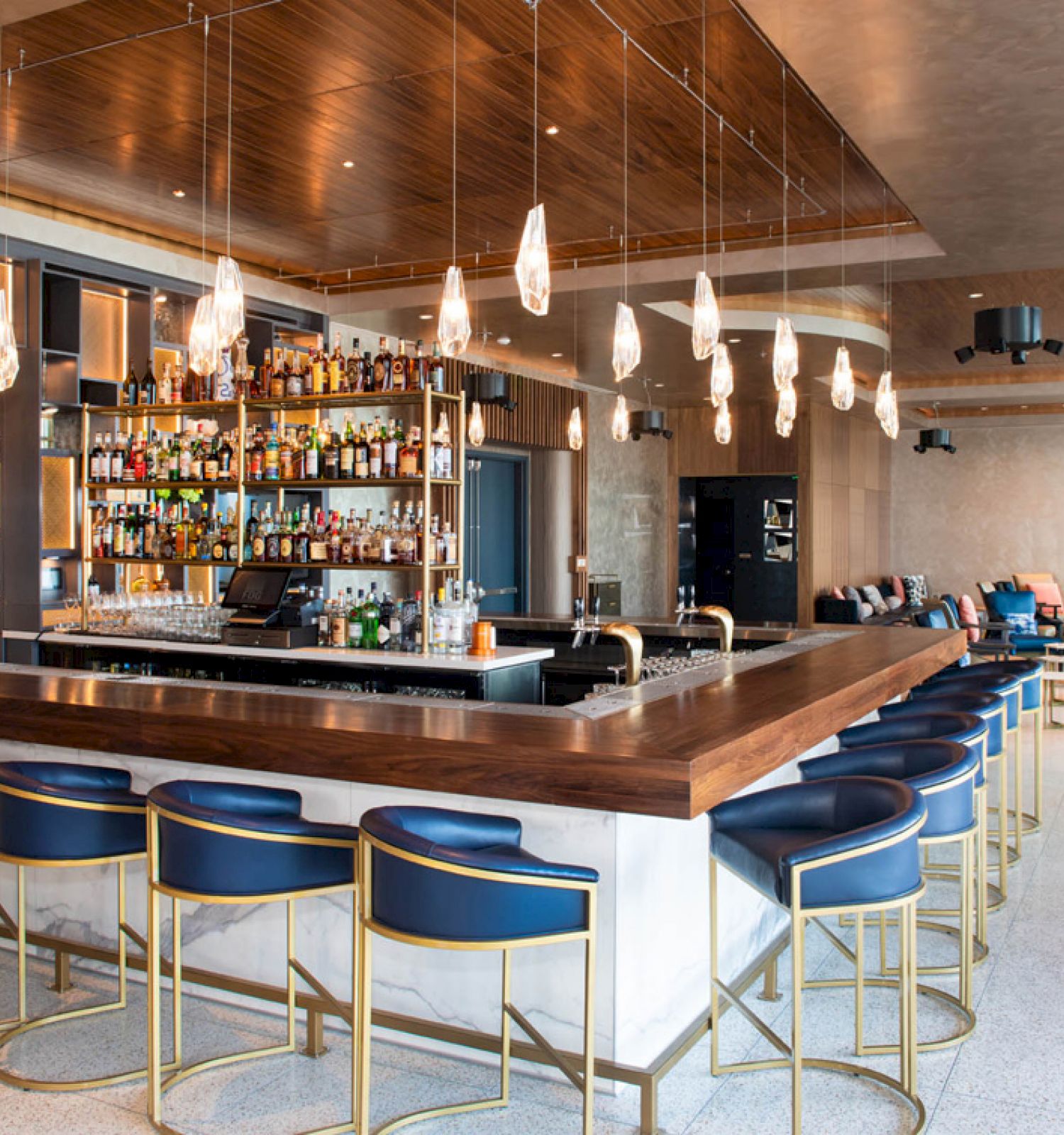 The image shows a modern bar with a wood and glass design, featuring a well-stocked liquor shelf, blue seating, and hanging lights.