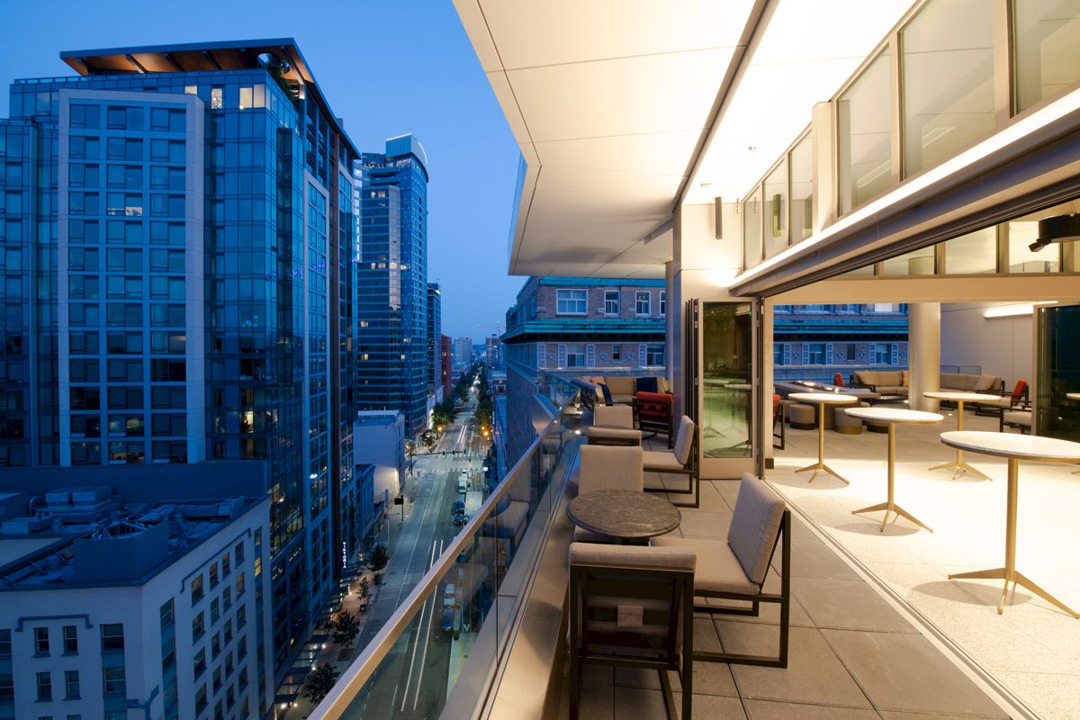 A modern balcony with tables and chairs overlooks a city street lined with tall, illuminated buildings at twilight.