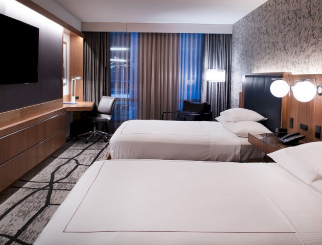 The image shows a modern hotel room with two beds, a TV, a desk with a chair, and stylish lighting and decor.