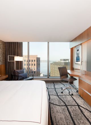 A modern hotel room with a large bed, desk, chair, and a flat-screen TV, offering a cityscape view through large windows.