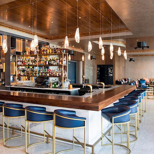 The image shows a modern bar with a central counter, blue chairs, hanging lights, and shelves of bottles, within a stylish lounge area.
