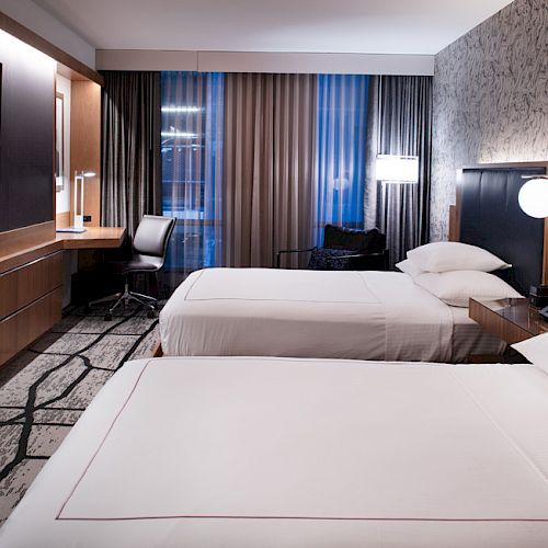 A modern hotel room features two beds, a large TV, a desk, and contemporary lighting and decor.