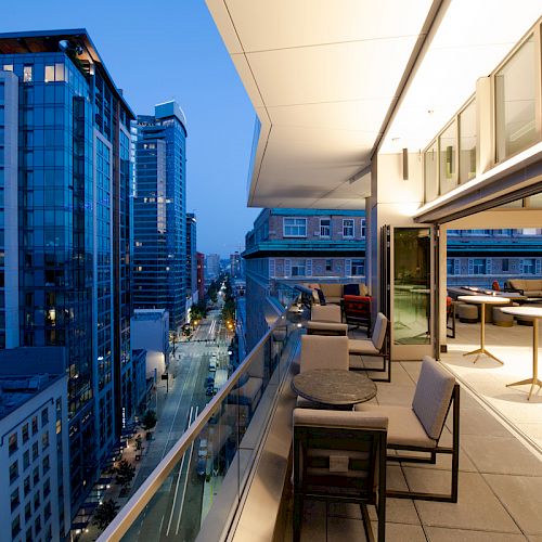 A modern cityscape with high-rise buildings and an elegant rooftop terrace featuring seating and tables under evening lighting.