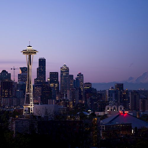 Seattle skyline at dusk with the Space Needle and Mount Rainier visible in the background, under a purple-blue sky.