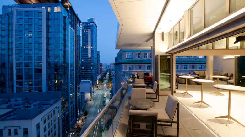A modern urban balcony overlooks city skyscrapers at dusk. It features seating areas with tables, illuminated by warm interior lighting.