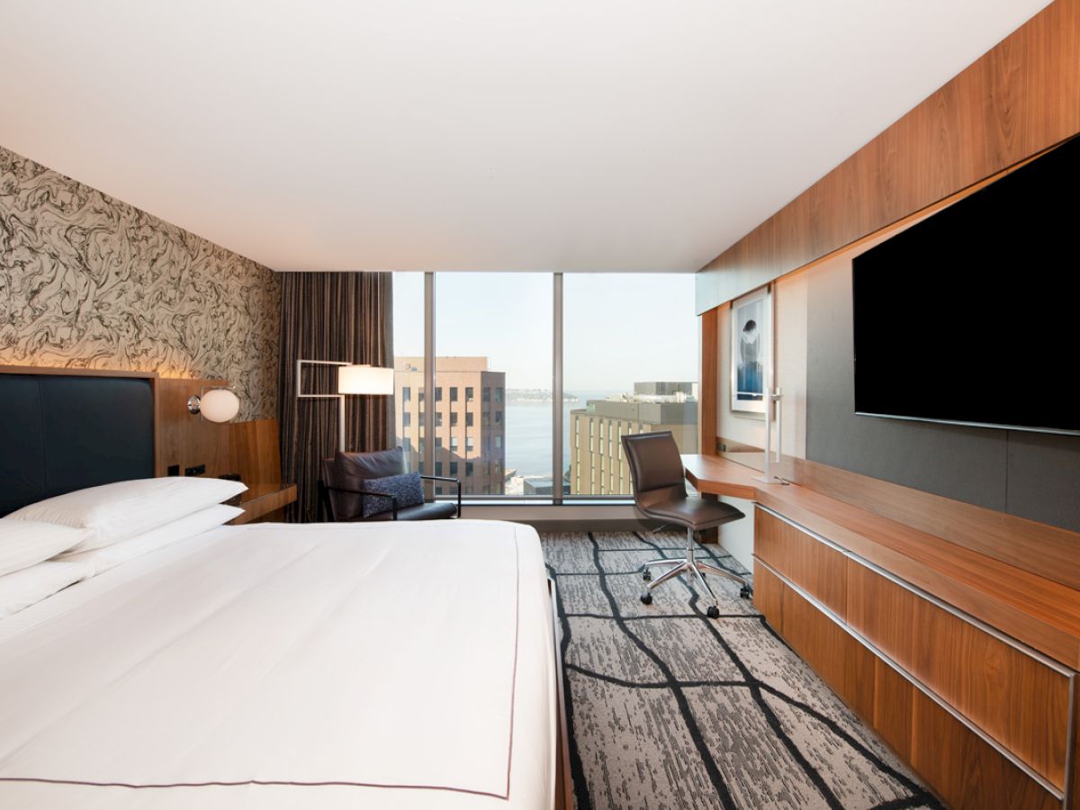 A modern hotel room with a large bed, desk, chair, TV, and a window view of city buildings and water.