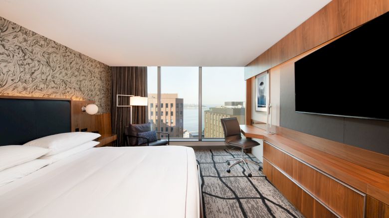 A modern hotel room with a large bed, desk, chair, TV, and a window view of city buildings and water.
