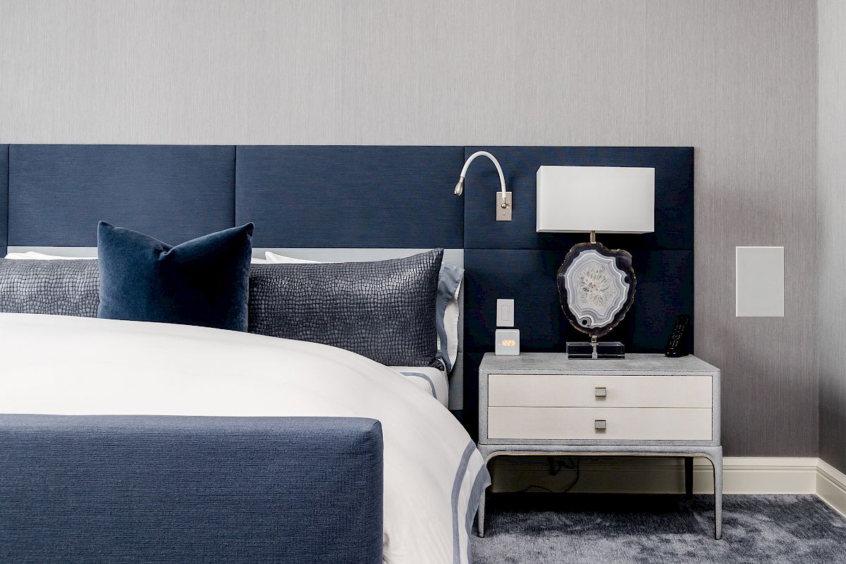 A modern bedroom with a blue headboard, white bed, and a nightstand with a lamp and decorative items on a carpeted floor.