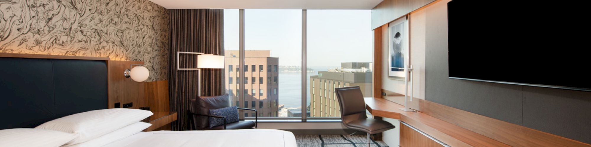 A modern hotel room with a large bed, desk, chair, and TV, featuring a cityscape view through the window.