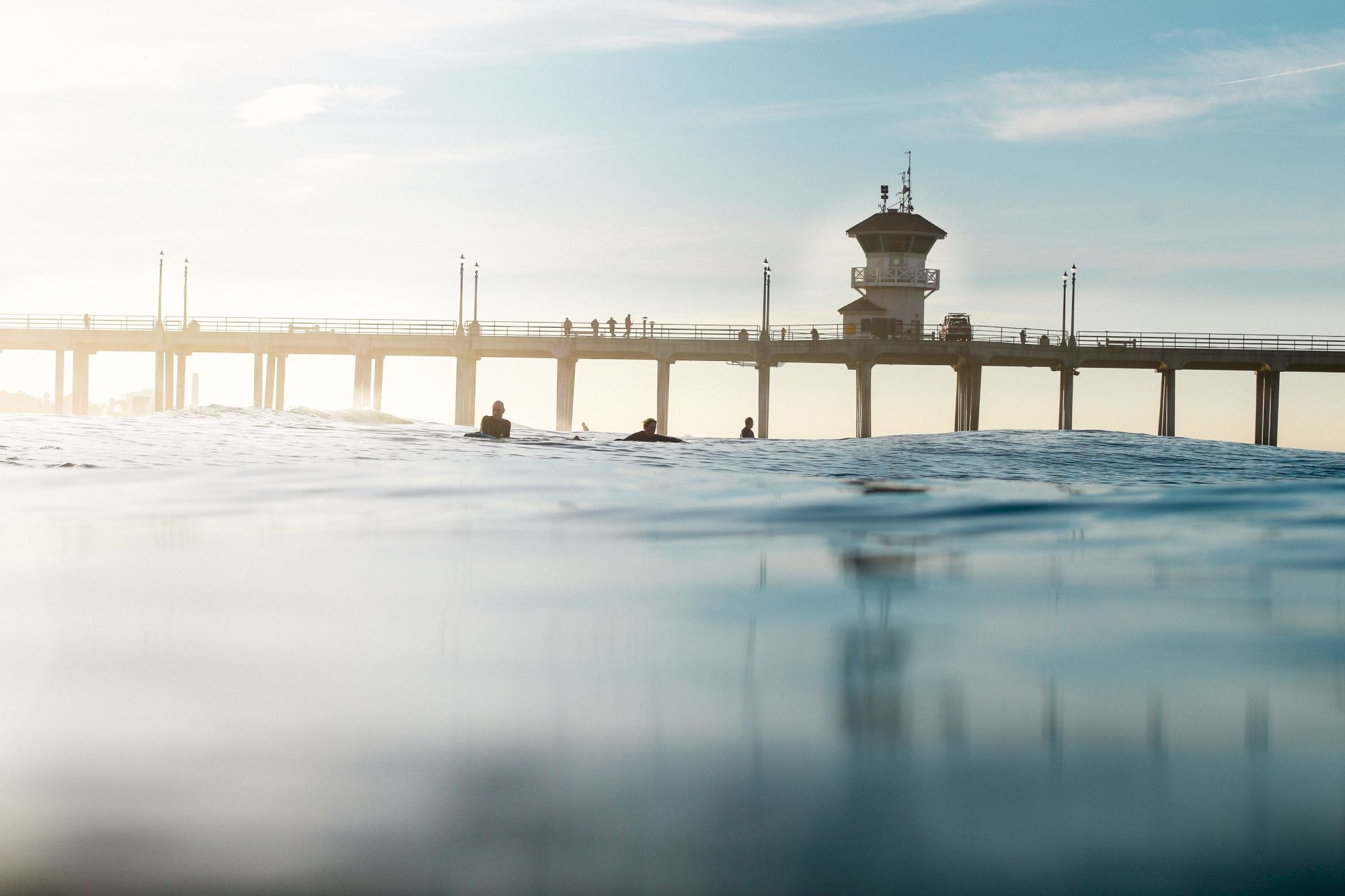 A pier stretches across the water with silhouettes of people. The scene includes a tranquil sky and gentle waves in the foreground.